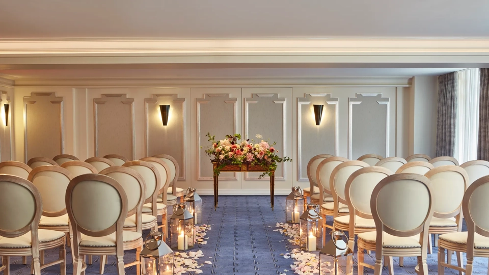 Elegant wedding ceremony setup at Claridge’s with cream chairs arranged in rows, an aisle lined with candles and flower petals, and a floral arrangement displayed on a wooden table at the front.