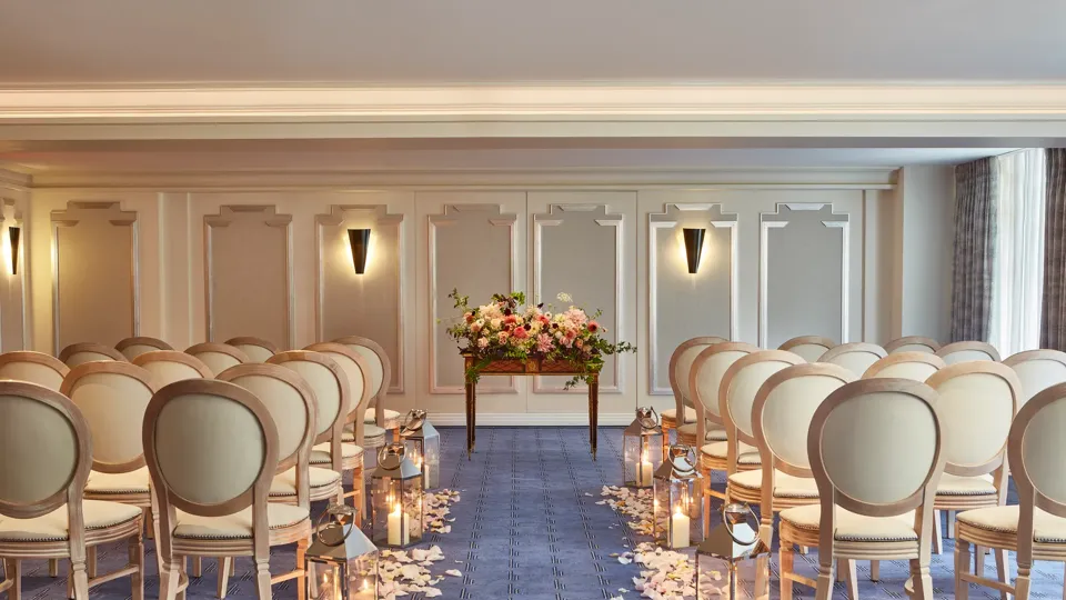 Elegant wedding ceremony setup at Claridge’s with cream chairs arranged in rows, an aisle lined with candles and flower petals, and a floral arrangement displayed on a wooden table at the front.