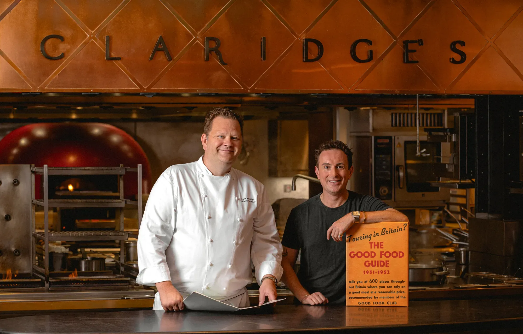 Two men standing at a kitchen counter beneath a copper sign reading “Claridge’s,” one wearing a white chef’s jacket and the other in a black shirt holding a vintage “Good Food Guide” sign, with a professional kitchen in the background.