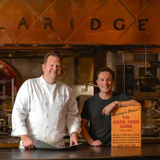 Two men standing at a kitchen counter beneath a copper sign reading “Claridge’s,” one wearing a white chef’s jacket and the other in a black shirt holding a vintage “Good Food Guide” sign, with a professional kitchen in the background.