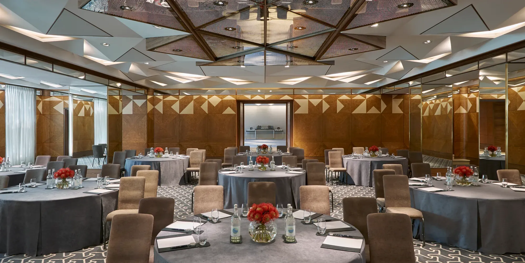 Conference room with round tables, grey tablecloths, notepads, water bottles, and red floral centerpieces under geometric mirrored ceiling.