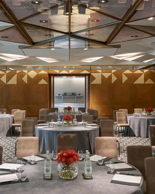 Conference room with round tables, grey tablecloths, notepads, water bottles, and red floral centerpieces under geometric mirrored ceiling.