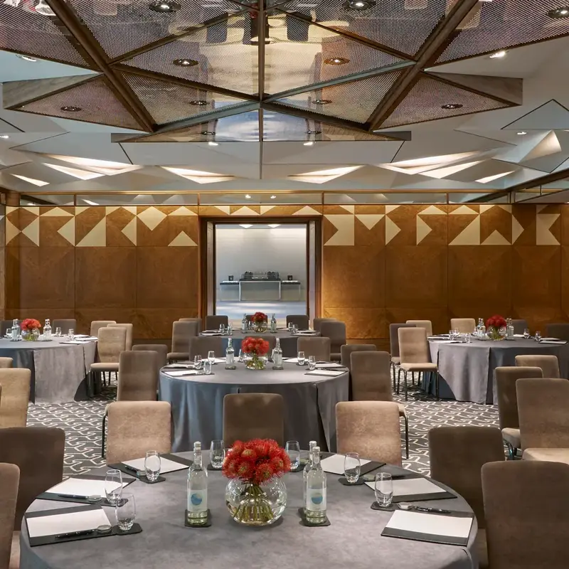 Conference room with round tables, grey tablecloths, notepads, water bottles, and red floral centerpieces under geometric mirrored ceiling.