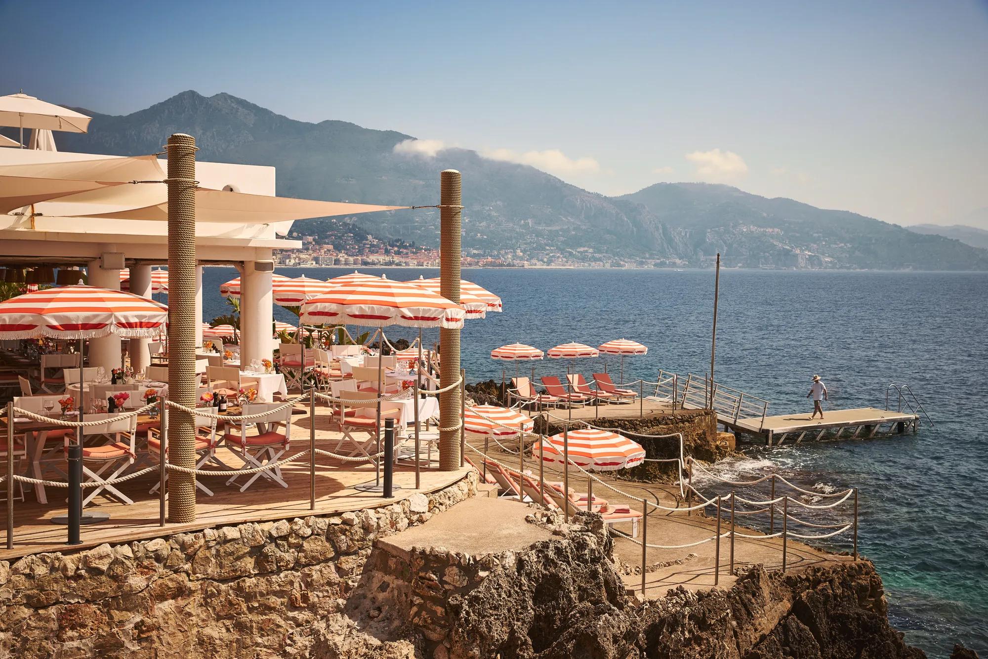 Seaside restaurant terrace with red-and-white umbrellas, dining tables, and sun loungers on rocky shoreline with mountain views.