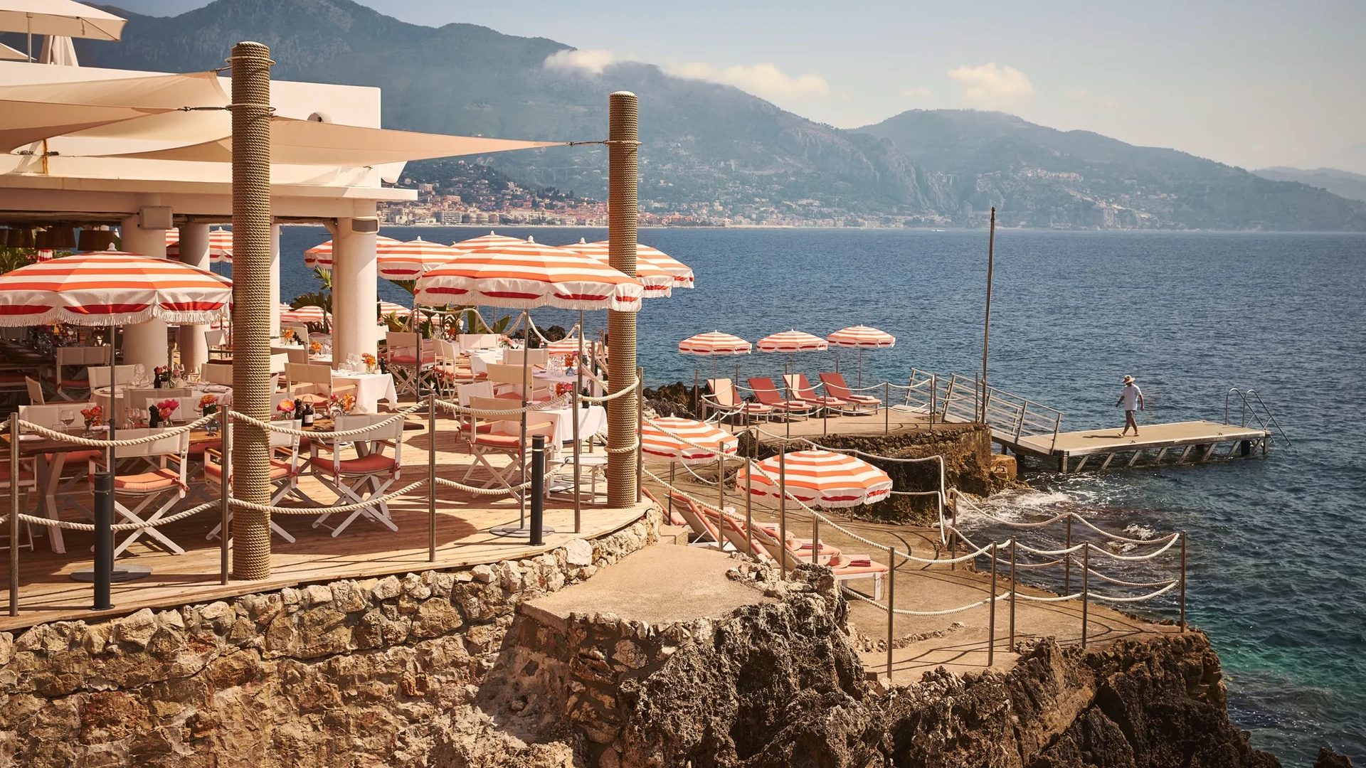 Seaside restaurant terrace with red-and-white umbrellas, dining tables, and sun loungers on rocky shoreline with mountain views.