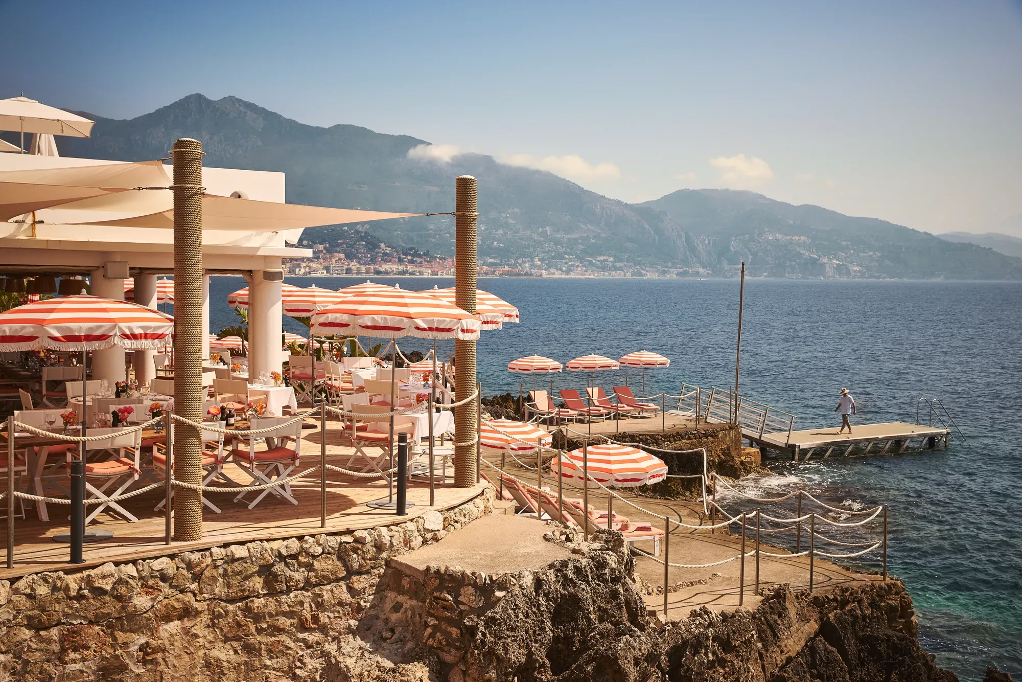 Seaside restaurant terrace with red-and-white umbrellas, dining tables, and sun loungers on rocky shoreline with mountain views.