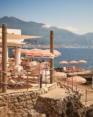 Seaside restaurant terrace with red-and-white umbrellas, dining tables, and sun loungers on rocky shoreline with mountain views.