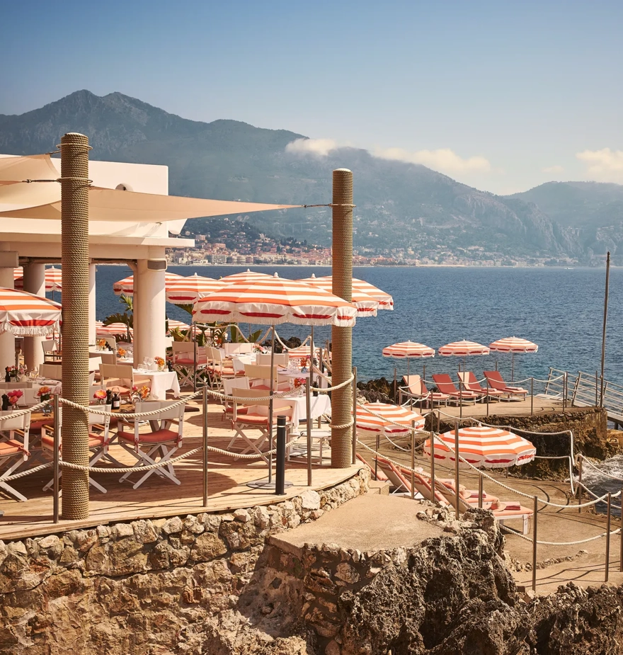 Seaside restaurant terrace with red-and-white umbrellas, dining tables, and sun loungers on rocky shoreline with mountain views.