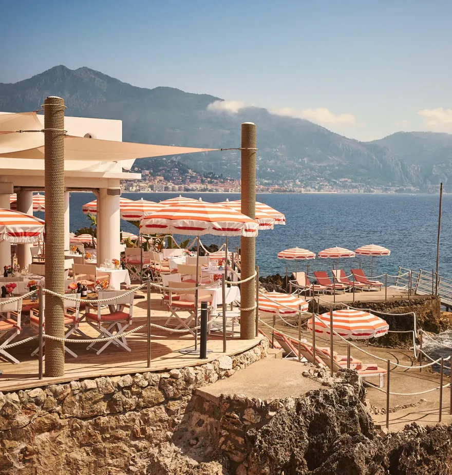 Seaside restaurant terrace with red-and-white umbrellas, dining tables, and sun loungers on rocky shoreline with mountain views.