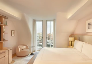 Light-filled bedroom with curved walls, a king-size bed, and glass doors opening onto a balcony with city views, furnished in soft neutral tones.