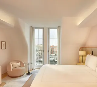 Light-filled bedroom with curved walls, a king-size bed, and glass doors opening onto a balcony with city views, furnished in soft neutral tones.