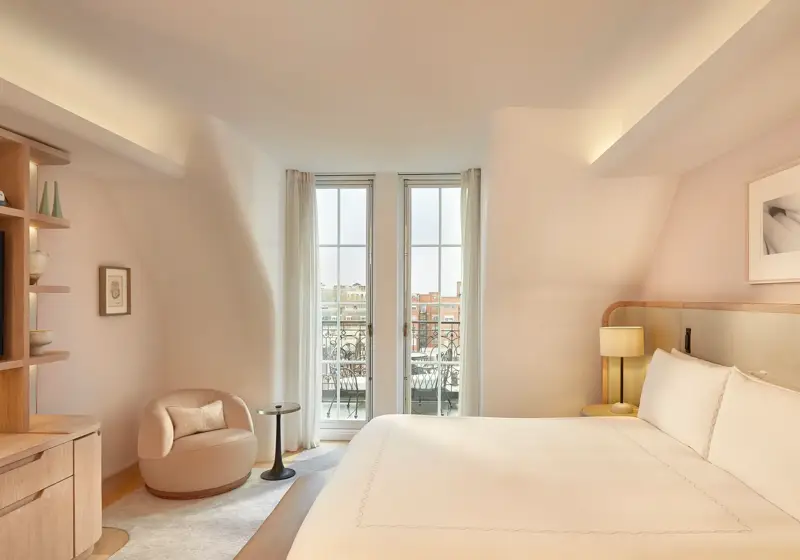 Light-filled bedroom with curved walls, a king-size bed, and glass doors opening onto a balcony with city views, furnished in soft neutral tones.