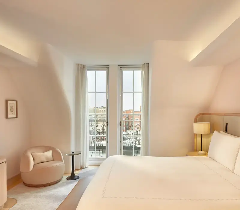 Light-filled bedroom with curved walls, a king-size bed, and glass doors opening onto a balcony with city views, furnished in soft neutral tones.