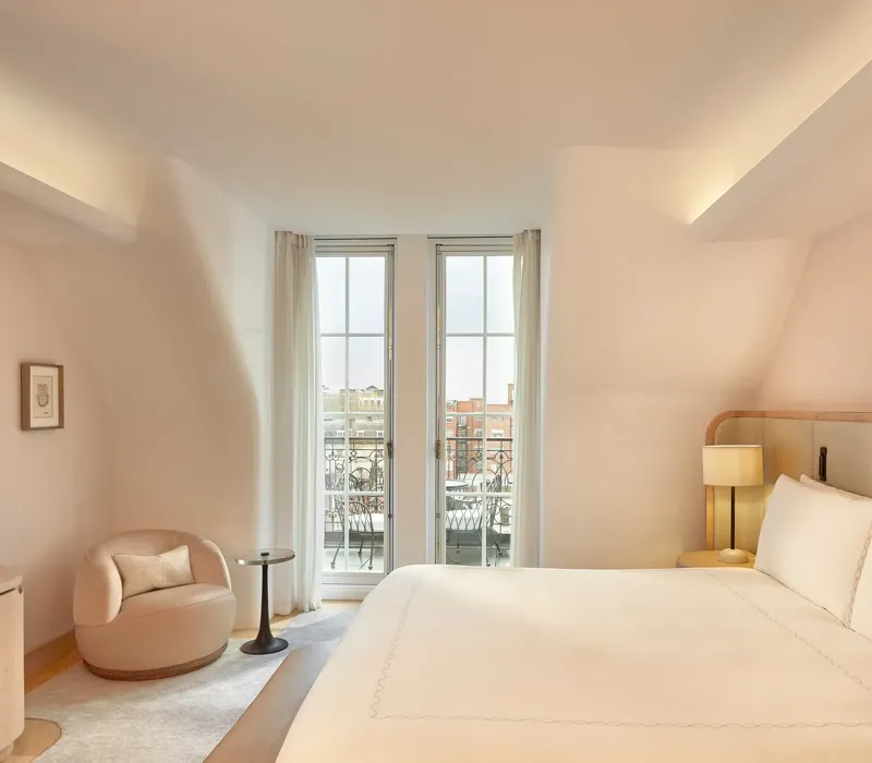 Light-filled bedroom with curved walls, a king-size bed, and glass doors opening onto a balcony with city views, furnished in soft neutral tones.