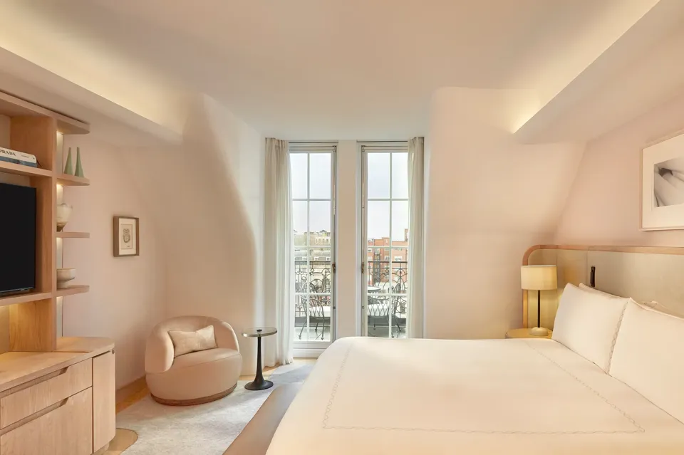 Light-filled bedroom with curved walls, a king-size bed, and glass doors opening onto a balcony with city views, furnished in soft neutral tones.