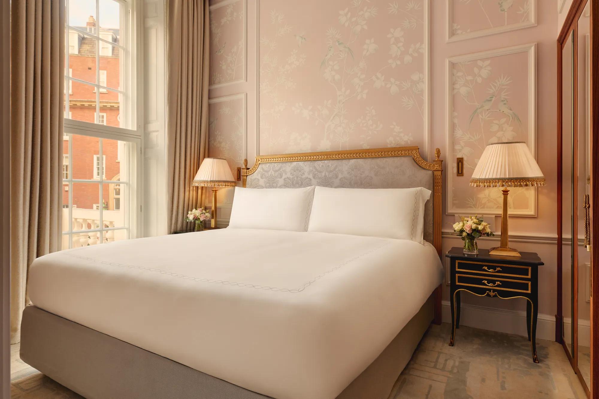 Elegant bedroom with king bed, pink floral walls, bedside lamps, and tall window.