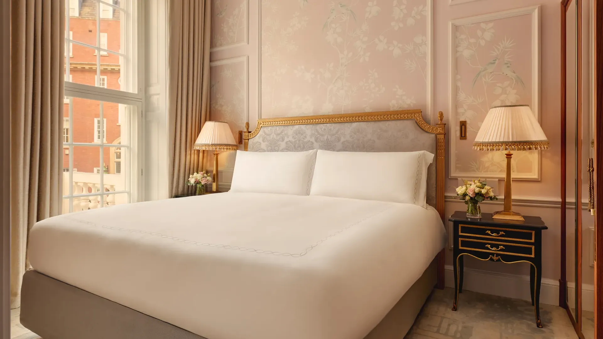 Elegant bedroom with king bed, pink floral walls, bedside lamps, and tall window.