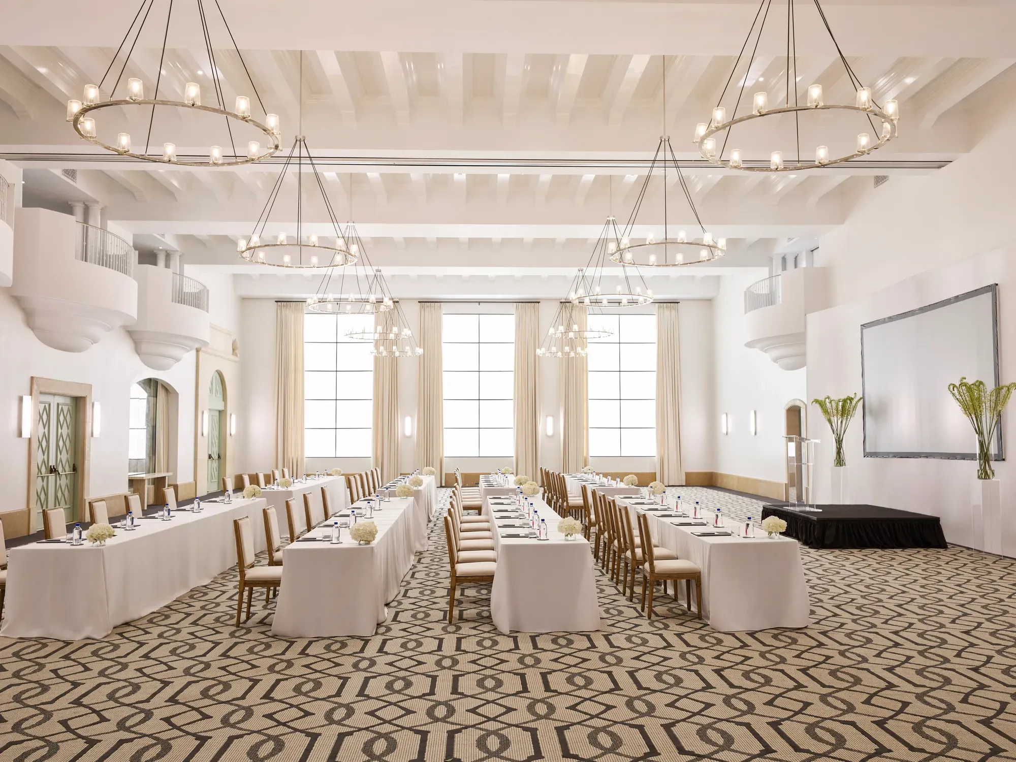 Grand ballroom in classroom conference setup with long draped tables, chandeliers, tall windows, and a presentation screen.