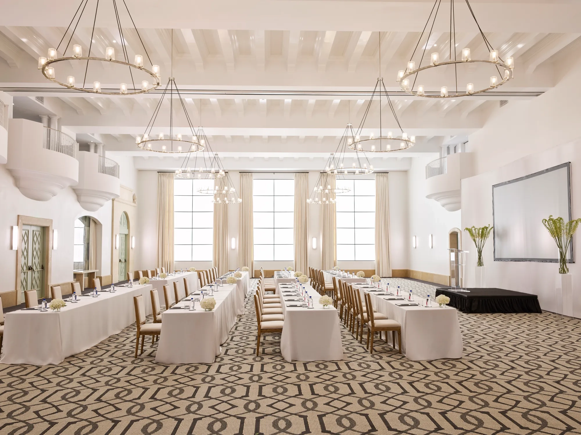 Grand ballroom in classroom conference setup with long draped tables, chandeliers, tall windows, and a presentation screen.