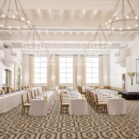 Grand ballroom in classroom conference setup with long draped tables, chandeliers, tall windows, and a presentation screen.