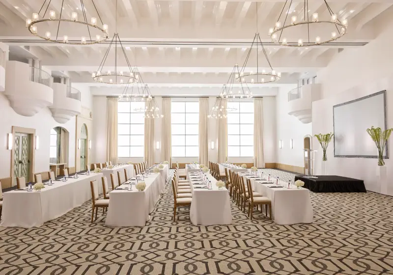 Grand ballroom in classroom conference setup with long draped tables, chandeliers, tall windows, and a presentation screen.