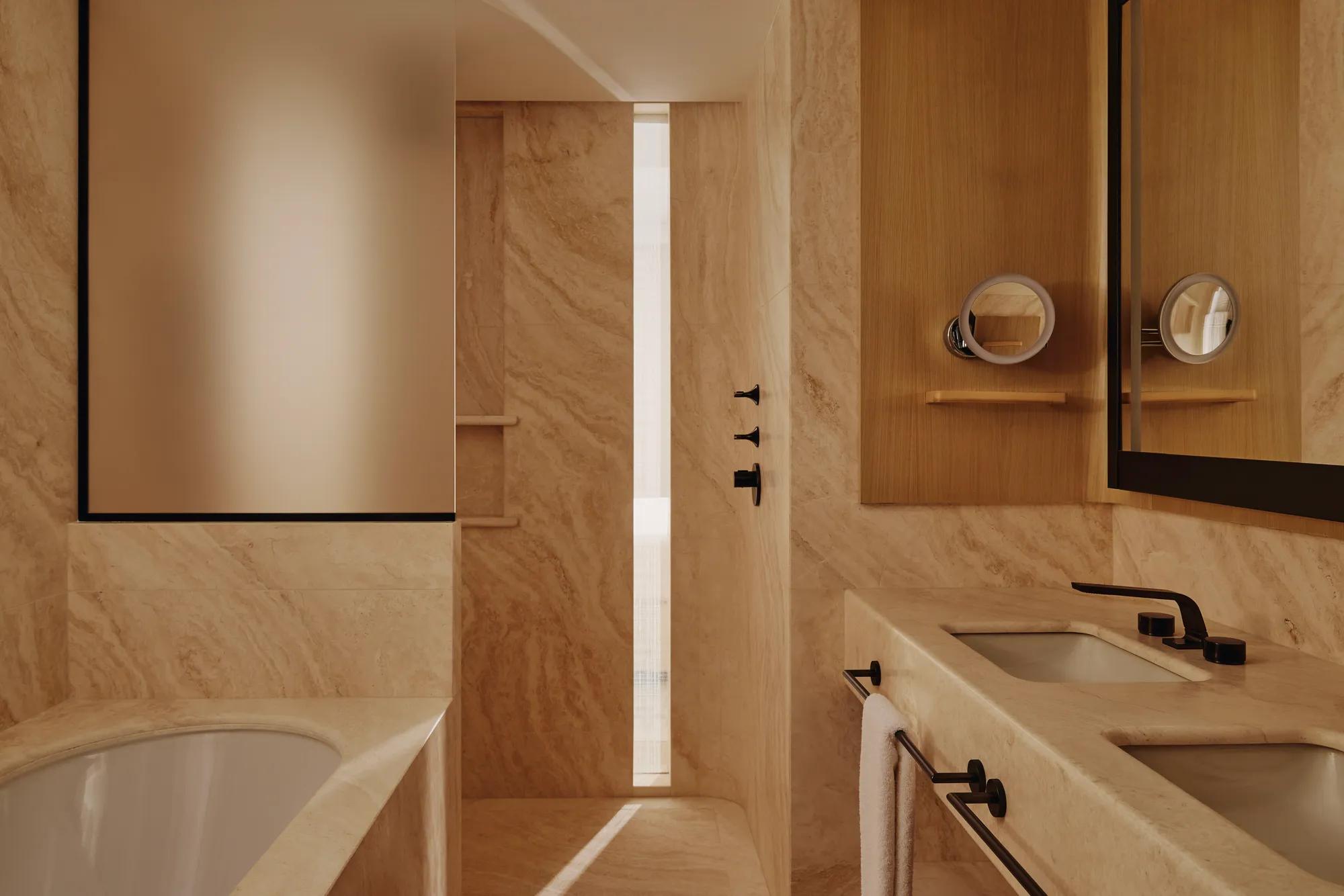 Luxury bathroom with marble walls, walk-in shower, freestanding bath edge, double vanity and warm wood cabinetry.