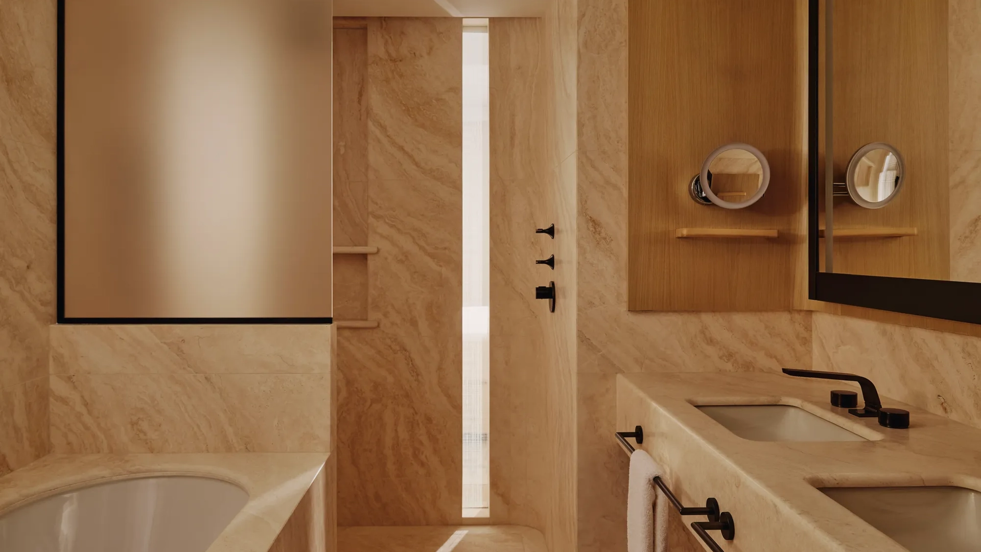 Luxury bathroom with marble walls, walk-in shower, freestanding bath edge, double vanity and warm wood cabinetry.