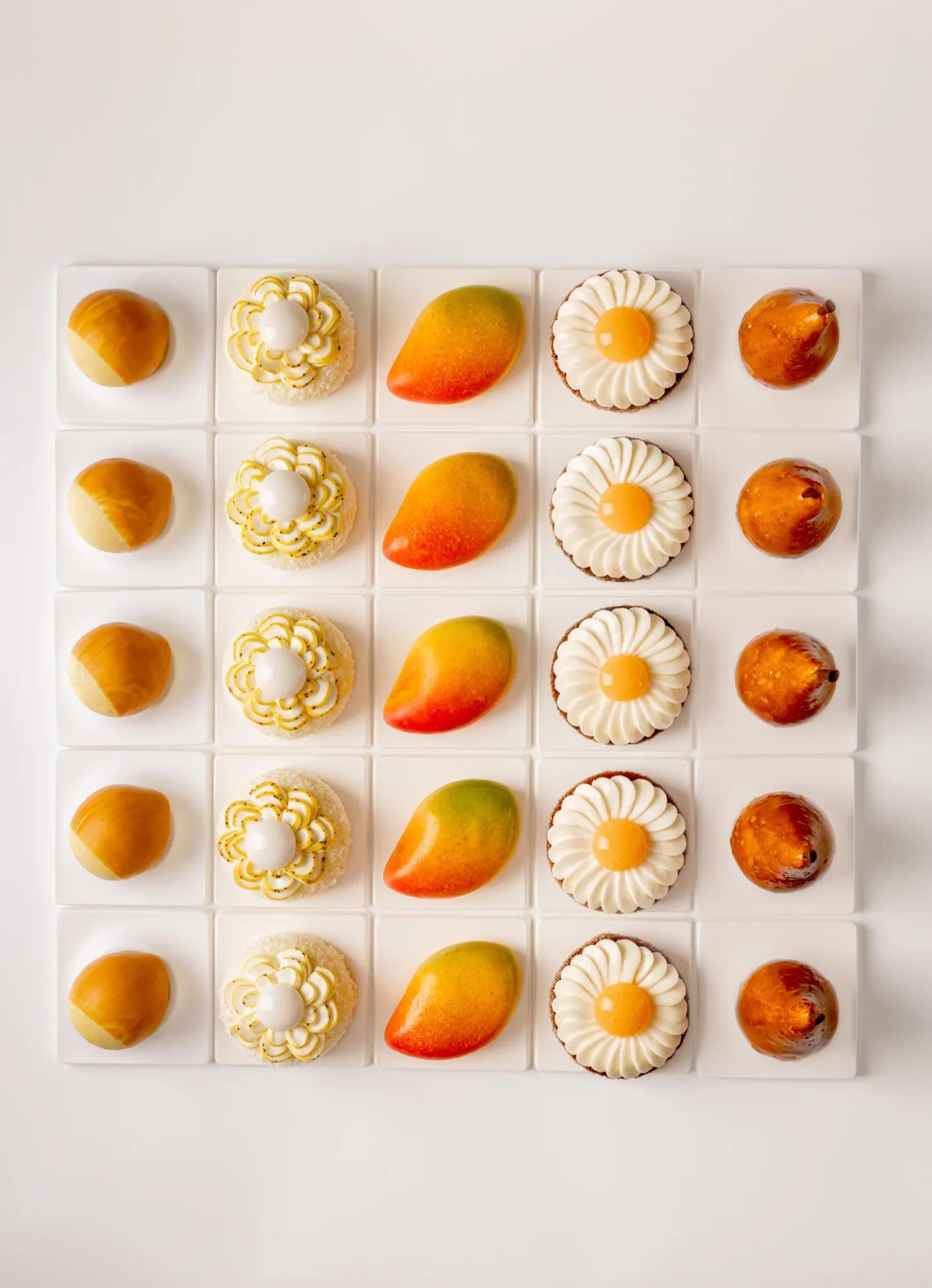 Five rows of miniature pastries including mango, pear and floral cakes, arranged in a neat grid on white background