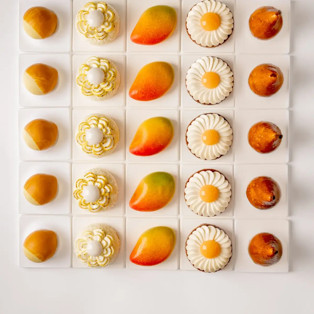 Five rows of miniature pastries including mango, pear and floral cakes, arranged in a neat grid on white background
