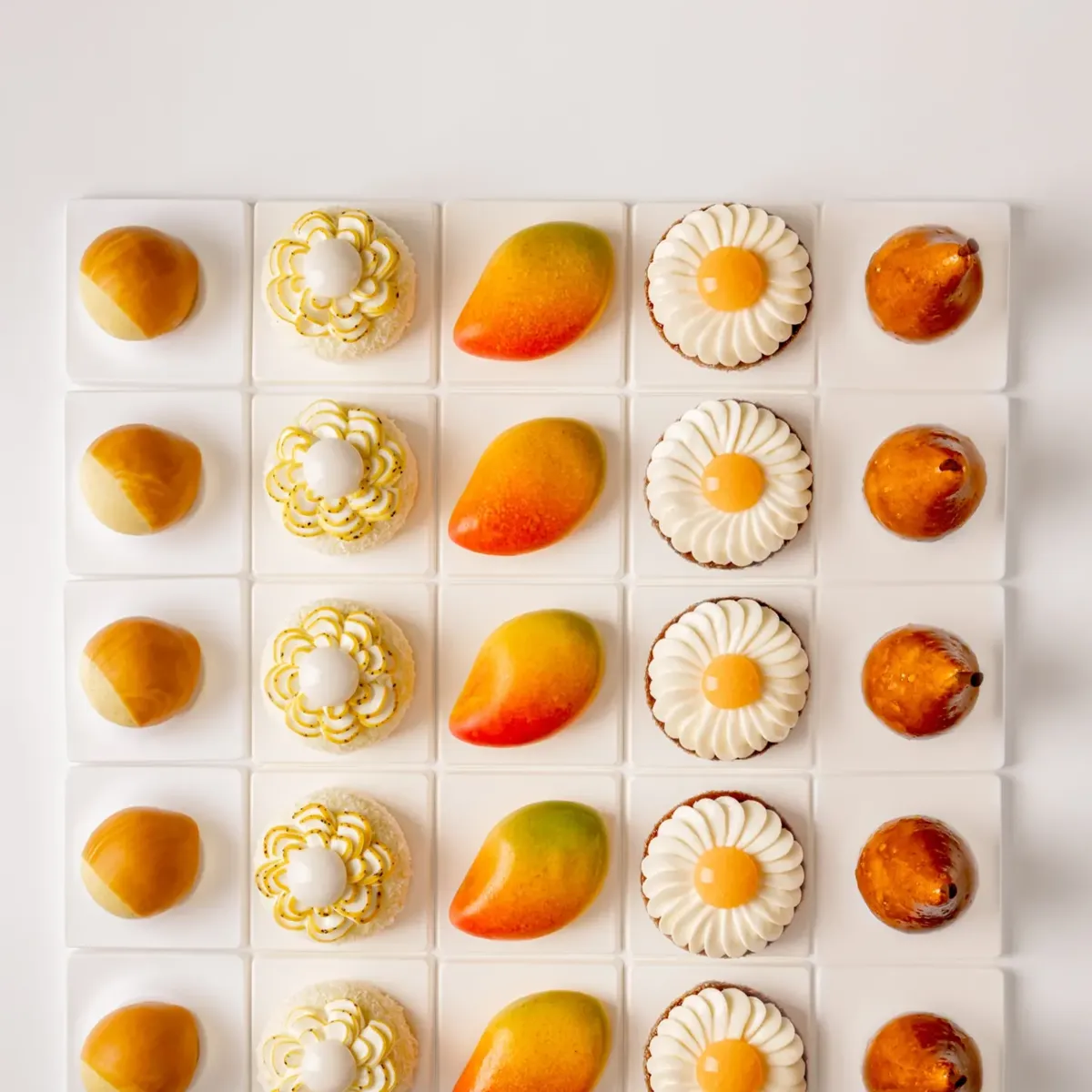 Five rows of miniature pastries including mango, pear and floral cakes, arranged in a neat grid on white background