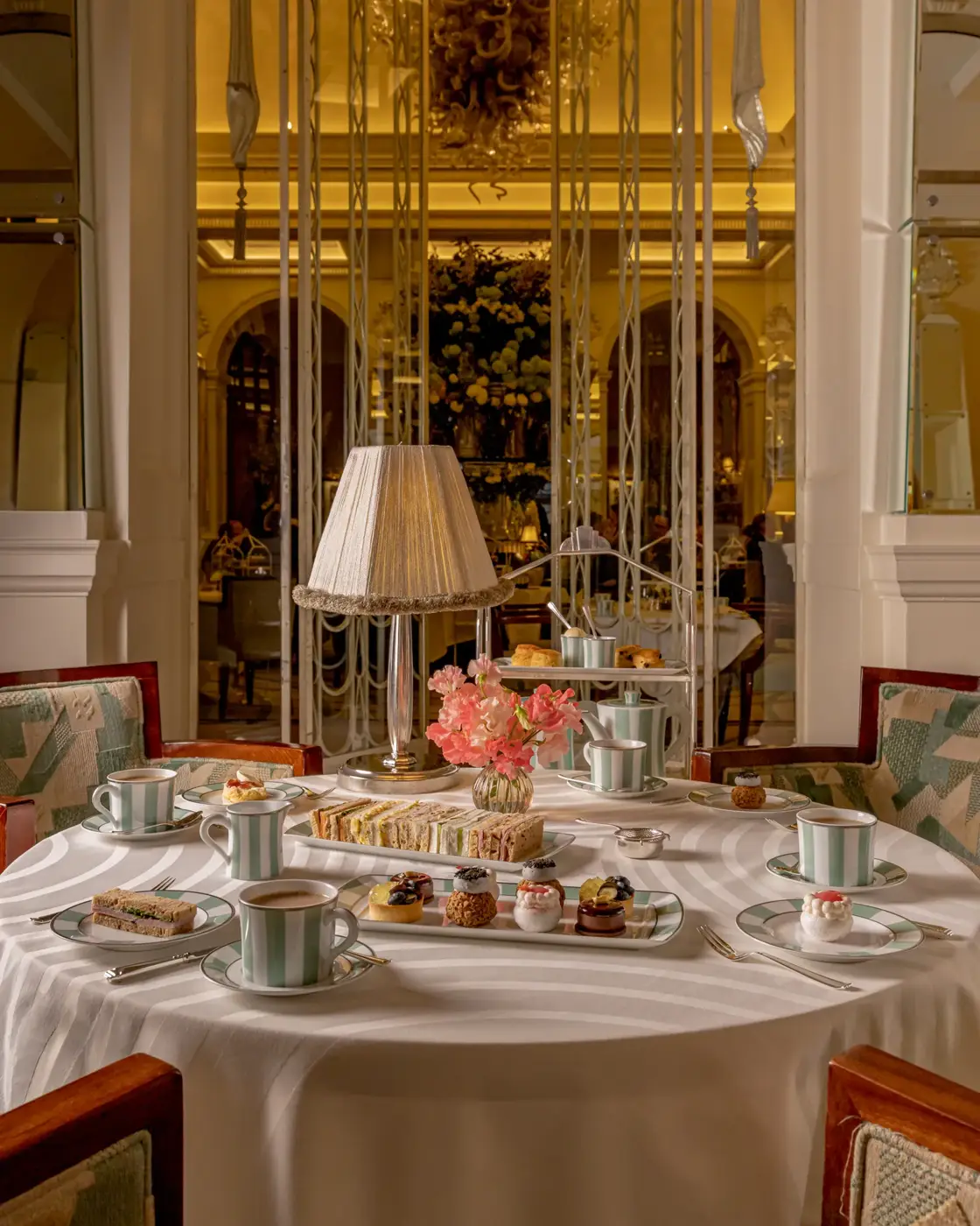 Afternoon tea table with sandwiches, pastries and striped teacups beneath a lamp in an ornate golden dining room.