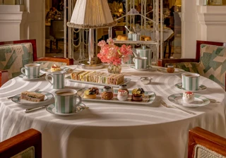 Afternoon tea table with sandwiches, pastries and striped teacups beneath a lamp in an ornate golden dining room.