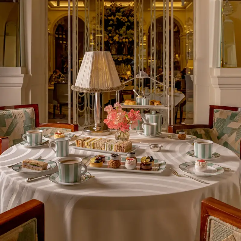 Afternoon tea table with sandwiches, pastries and striped teacups beneath a lamp in an ornate golden dining room.