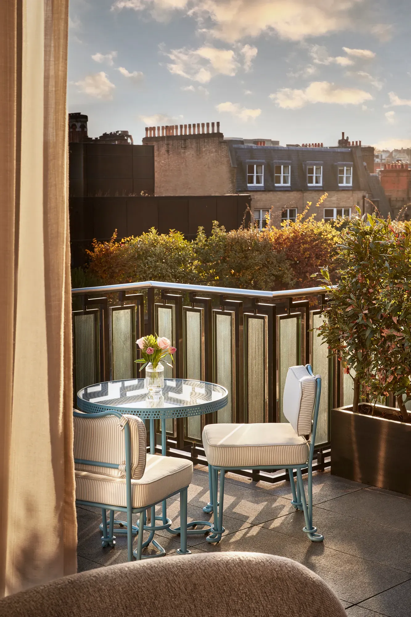 Private terrace with a small blue bistro table and two chairs overlooking rooftops and greenery, with soft evening light and a vase of flowers on the table.