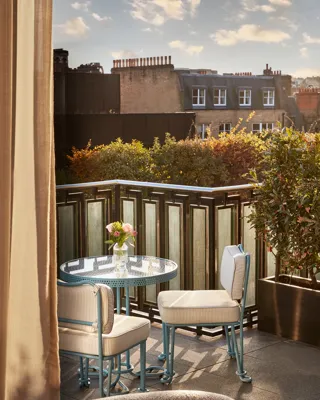 Private terrace with a small blue bistro table and two chairs overlooking rooftops and greenery, with soft evening light and a vase of flowers on the table.