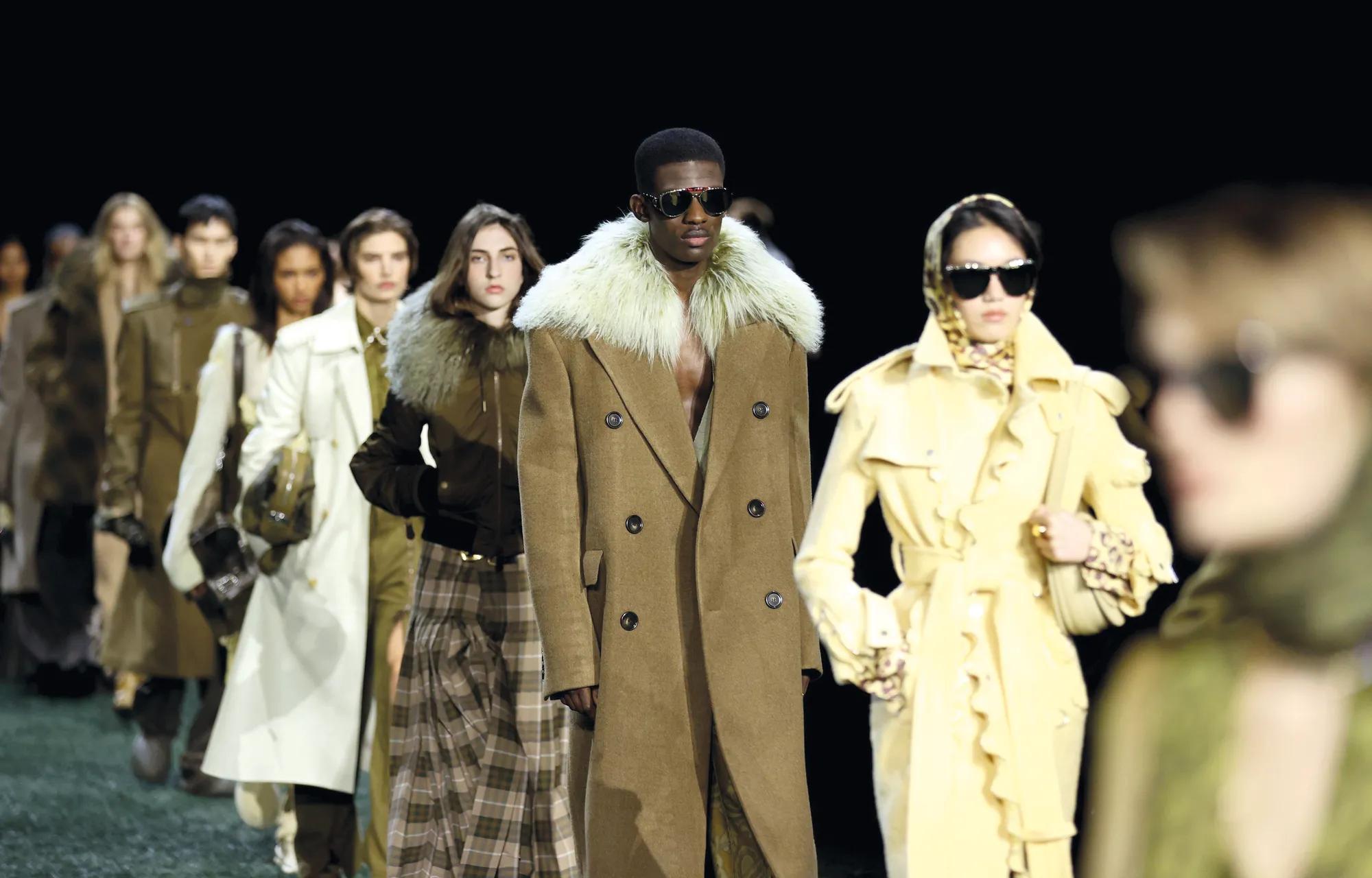 Models walk in formation on a fashion runway wearing tailored coats in neutral tones.