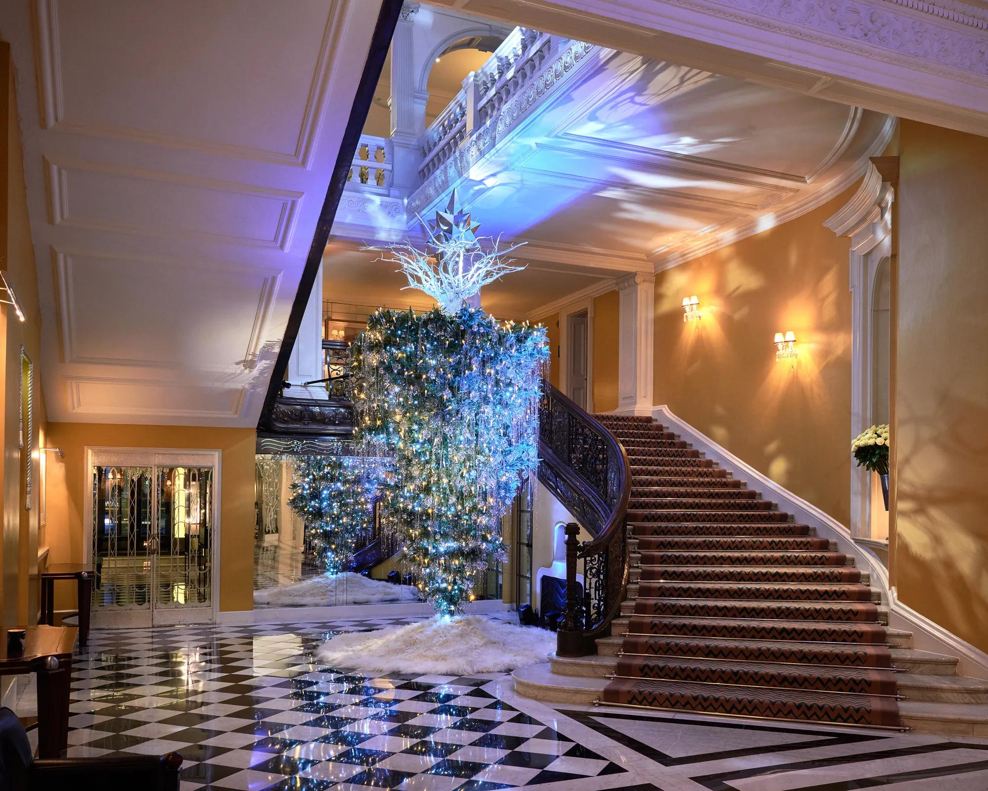 Grand hotel lobby with sweeping staircase and checkered floor, featuring illuminated cascading tree installation with blue lighting and star detail above.