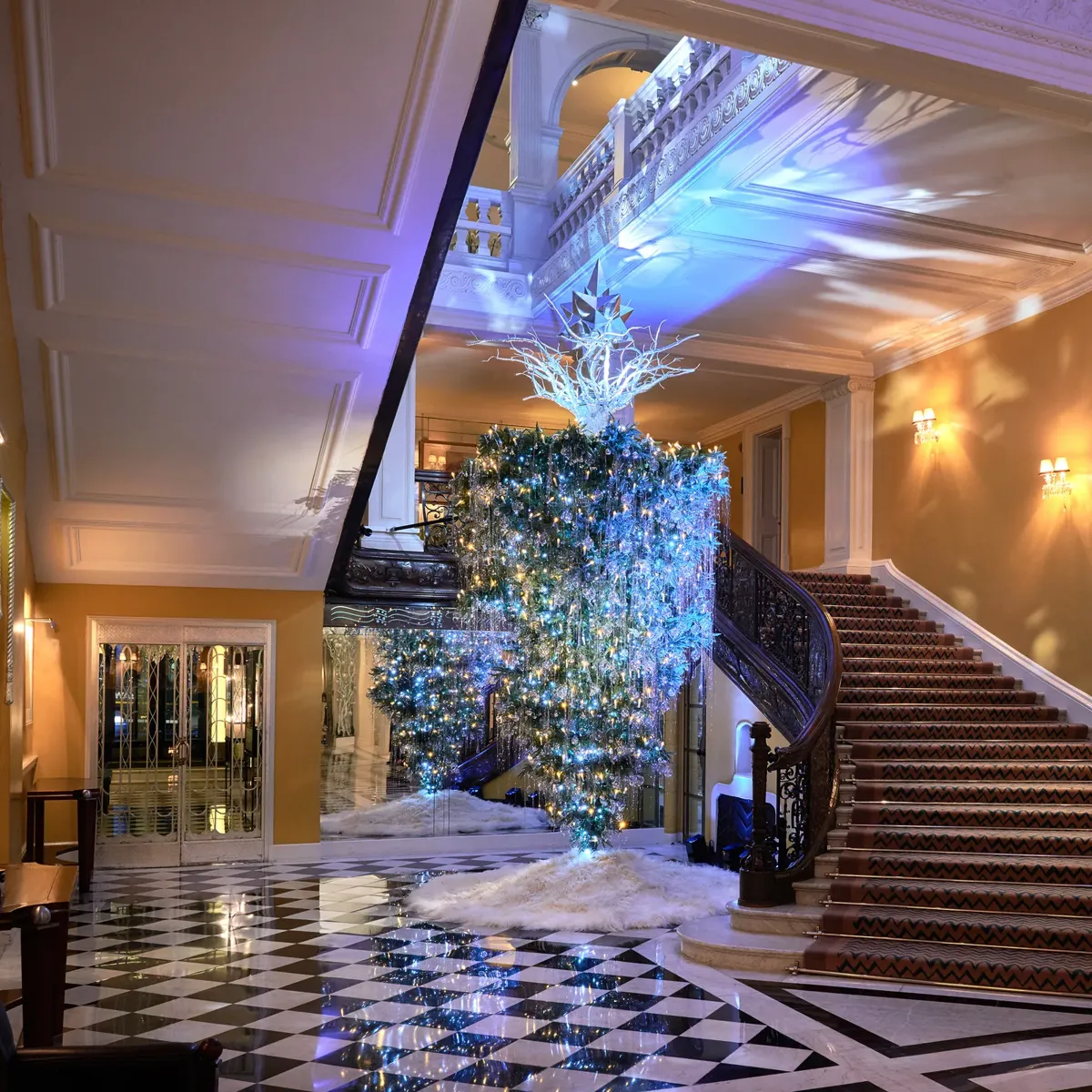 Grand hotel lobby with sweeping staircase and checkered floor, featuring illuminated cascading tree installation with blue lighting and star detail above.