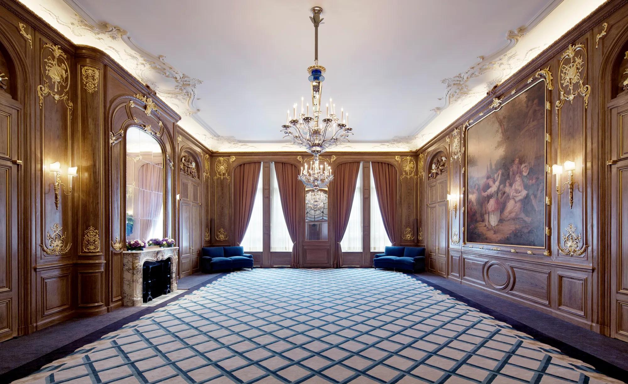 Spacious Claridge’s ballroom with ornate wood-panelled walls, grand chandeliers, and a large classical painting, featuring deep blue sofas, floral displays, and tall curtained windows.