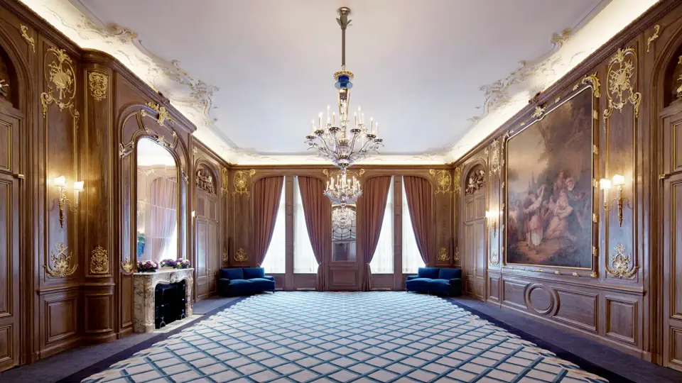 Spacious Claridge’s ballroom with ornate wood-panelled walls, grand chandeliers, and a large classical painting, featuring deep blue sofas, floral displays, and tall curtained windows.