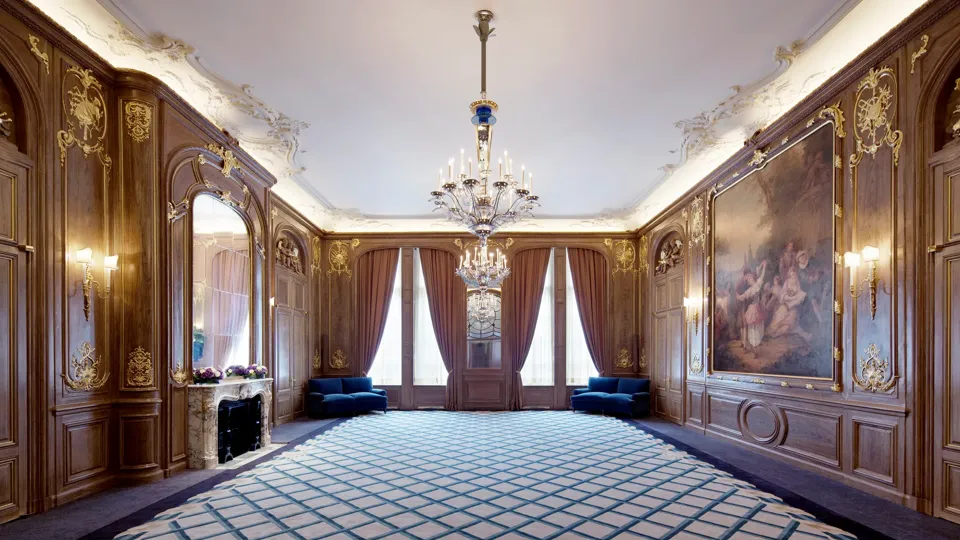 Spacious Claridge’s ballroom with ornate wood-panelled walls, grand chandeliers, and a large classical painting, featuring deep blue sofas, floral displays, and tall curtained windows.