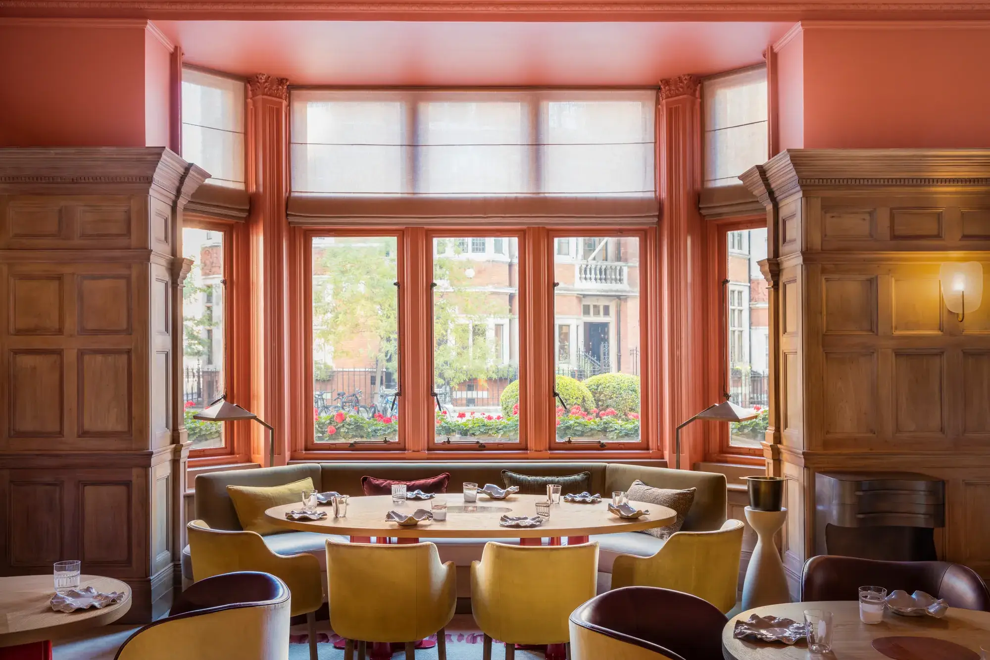 Bright dining room with coral walls, large bay windows, and colourful chairs arranged around round tables.