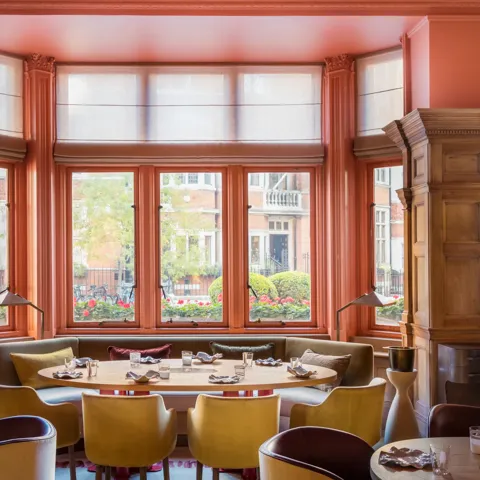 Bright dining room with coral walls, large bay windows, and colourful chairs arranged around round tables.
