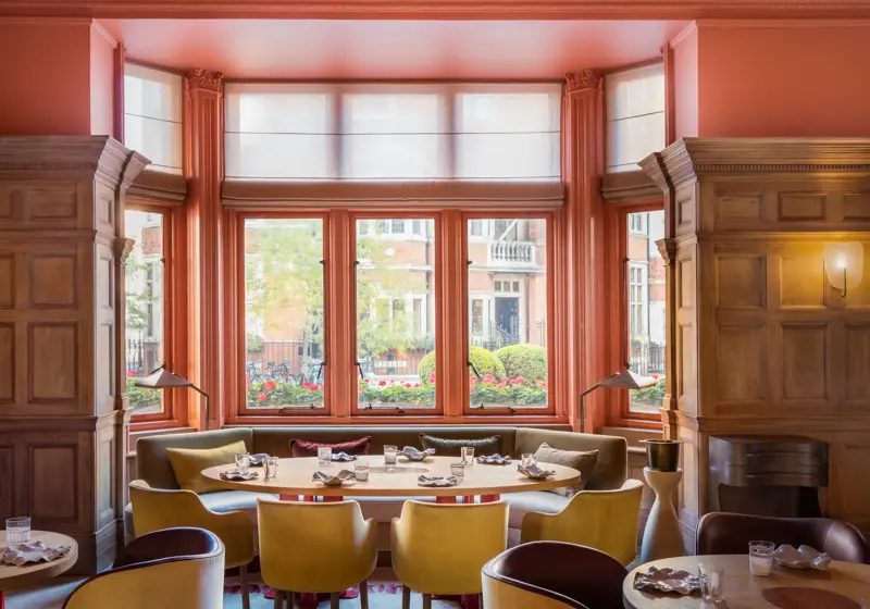 Bright dining room with coral walls, large bay windows, and colourful chairs arranged around round tables.
