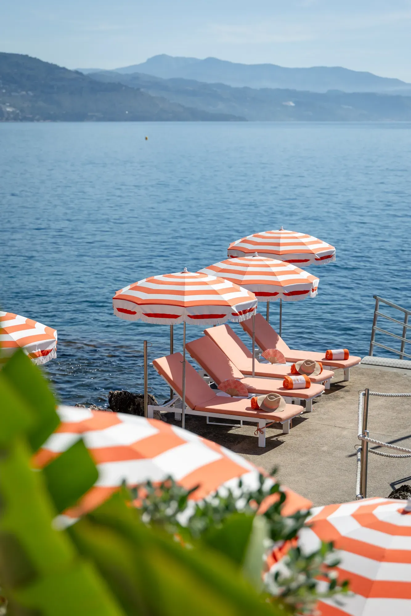 Striped orange-and-white umbrellas with sun loungers on a waterside terrace overlooking the sea.