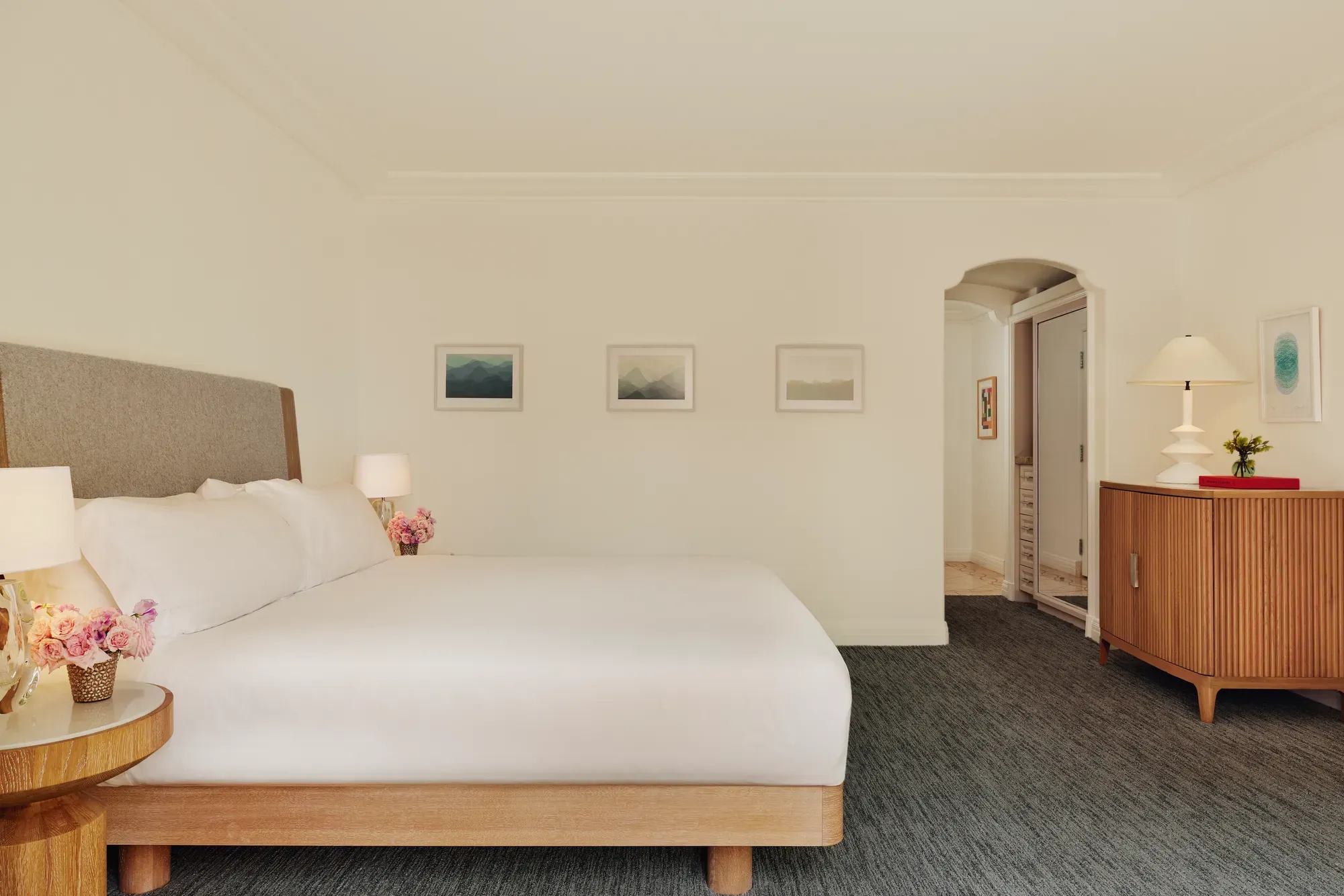 Accessible king bedroom with upholstered headboard, three framed prints, dresser with lamp, and an arched doorway to the bathroom.