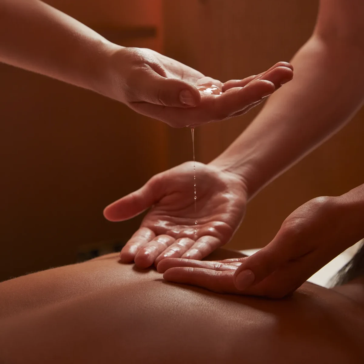 Close-up of two spa therapists pouring warm oil into hands during a treatment in softly lit, tranquil surroundings.