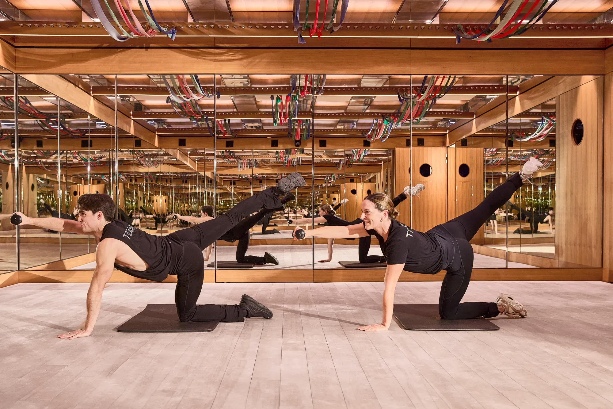 Two people performing floor exercises with weights on mats in mirrored studio, wooden interiors and ceiling cables visible.