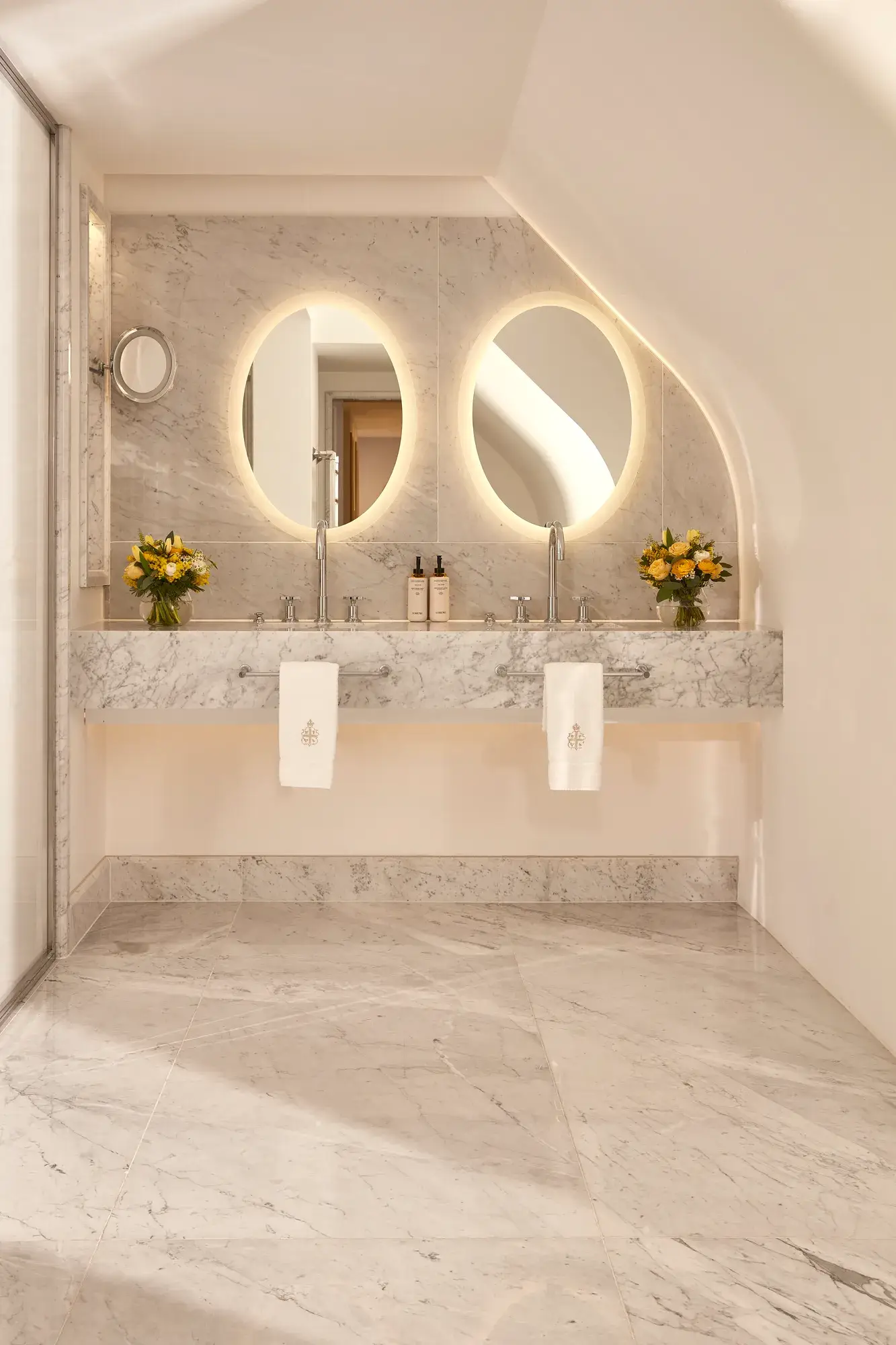 Elegant marble bathroom with twin sinks, circular backlit mirrors, and fresh yellow flower arrangements on either side of the vanity.