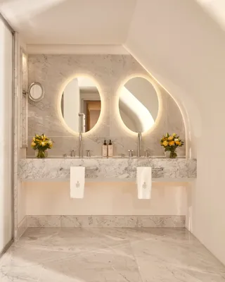 Elegant marble bathroom with twin sinks, circular backlit mirrors, and fresh yellow flower arrangements on either side of the vanity.